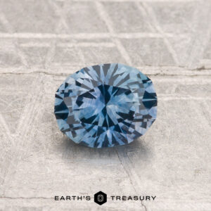 A 0.92-carat, round, faceted Montana sapphire (heated) is showcased on a textured metallic surface with "EARTH'S TREASURY" displayed at the bottom.