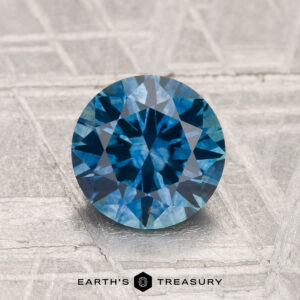 A 1.60-carat heated Montana Sapphire, round brilliant-cut and blue, is displayed on a textured gray surface with "EARTH'S TREASURY" at the bottom.