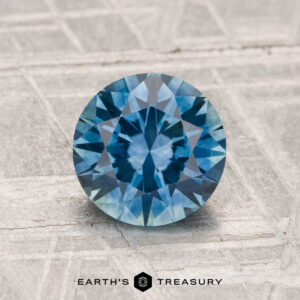 A 1.50-carat heated Montana sapphire gemstone is shown on a textured gray surface, with the Earth's Treasury logo at the bottom.