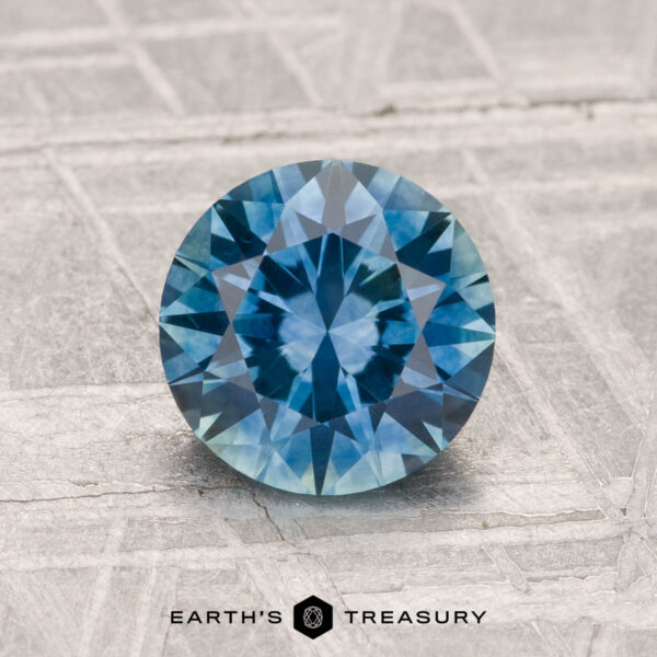 1.50-Carat Montana Sapphire (Heated) A 1.50-carat heated Montana sapphire gemstone is shown on a textured gray surface, with the Earth's Treasury logo at the bottom.
