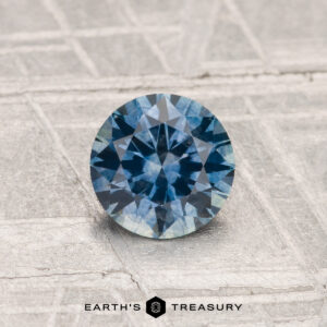A 0.82-Carat Montana Sapphire (Heated), round and faceted, is shown on a textured light gray surface with "EARTH'S TREASURY" at the bottom.