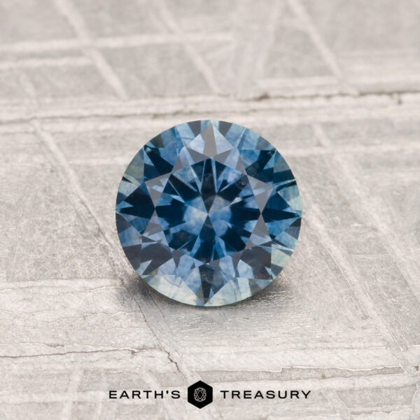 0.82-Carat Montana Sapphire (Heated) A 0.82-Carat Montana Sapphire (Heated), round and faceted, is shown on a textured light gray surface with "EARTH'S TREASURY" at the bottom.