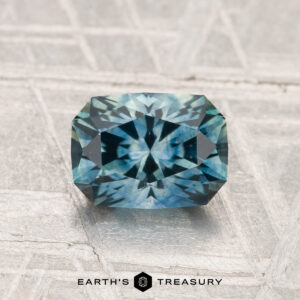 A 1.38-carat heated Montana sapphire with a rectangular, blue-green faceted cut is displayed on a textured light gray surface, with the "EARTH'S TREASURY" text and logo at the bottom.