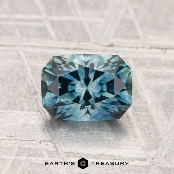 1.38-Carat Montana Sapphire (Heated) A 1.38-carat heated Montana sapphire with a rectangular, blue-green faceted cut is displayed on a textured light gray surface, with the "EARTH'S TREASURY" text and logo at the bottom.