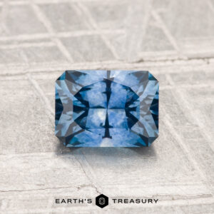 A 1.14-carat heated Montana sapphire with geometric faceting rests on a textured gray surface; the "Earth's Treasury" logo appears below the gemstone.