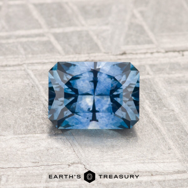 1.14-Carat Montana Sapphire (Heated) A 1.14-carat heated Montana sapphire with geometric faceting rests on a textured gray surface; the "Earth's Treasury" logo appears below the gemstone.