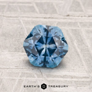 A faceted, hexagonal 1.12-carat heated Montana sapphire rests on a textured light gray surface, with "EARTH'S TREASURY" displayed at the bottom.