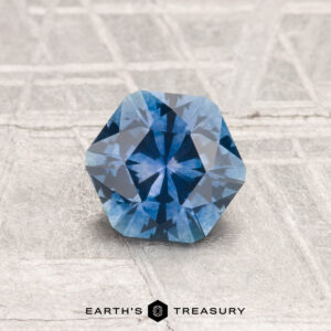 A faceted, hexagonal 0.97-carat heated Montana sapphire with a blue hue is displayed on a textured light gray surface featuring the Earth's Treasury logo at the bottom.