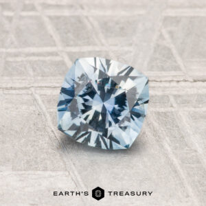A 1.00-carat heated Montana sapphire, cushion-cut and blue, is displayed on a textured gray surface with "EARTH'S TREASURY" at the bottom.