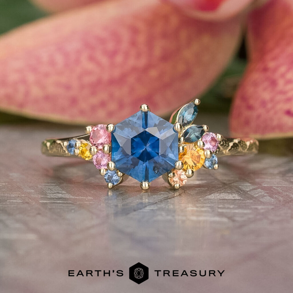 The eb7c0452-69dd-46c3-9196-7f43dfa2c8e5-1200-BW features a gold band with a hexagonal blue gemstone center, accented by small pink, yellow, and blue stones, showcased on a textured surface.