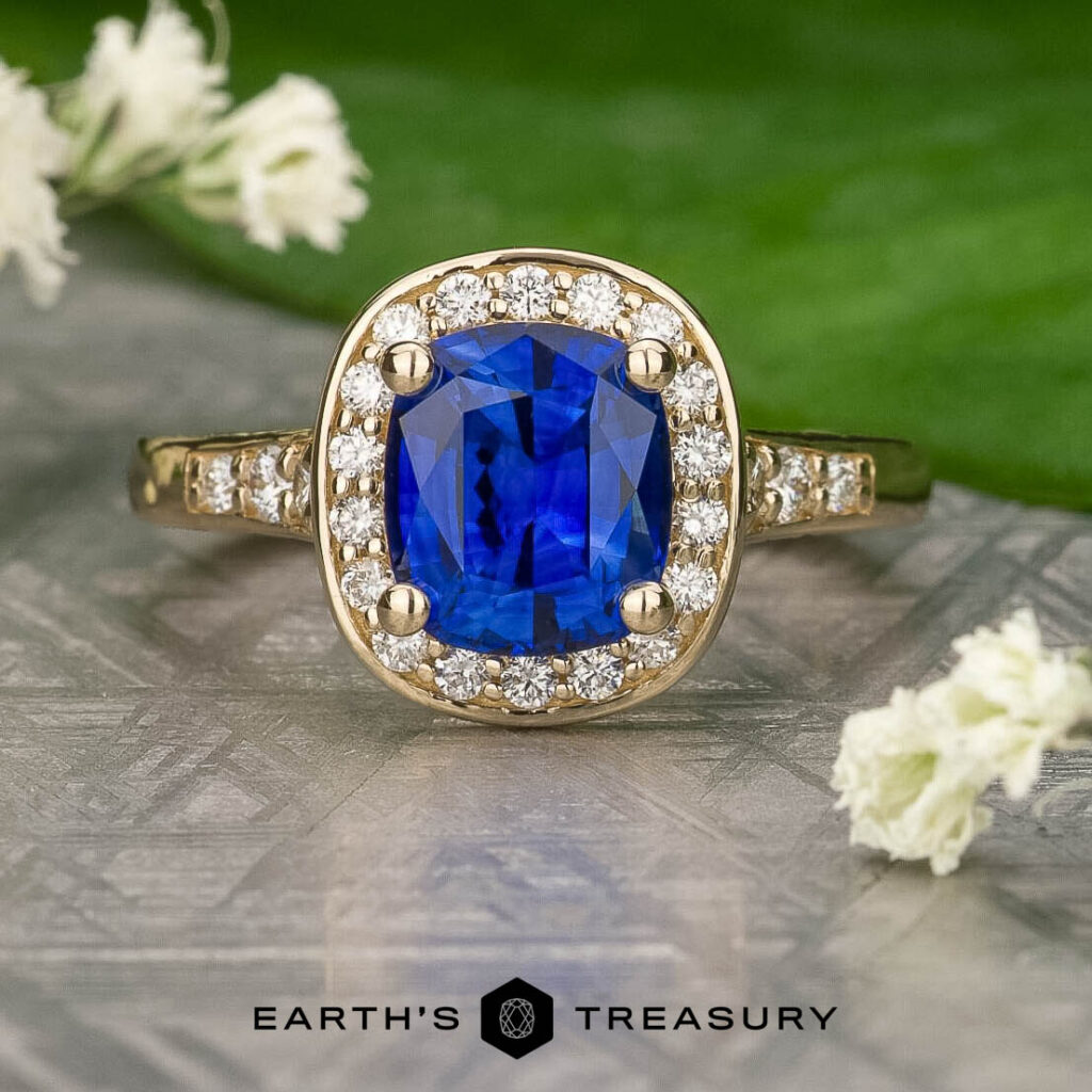 The f399e56b-9b79-46fd-bf3e-ea60d5241fb2-1200-BW gold ring features a cushion-cut blue sapphire center, accented by a diamond halo and band, displayed on a textured surface with small white flowers.