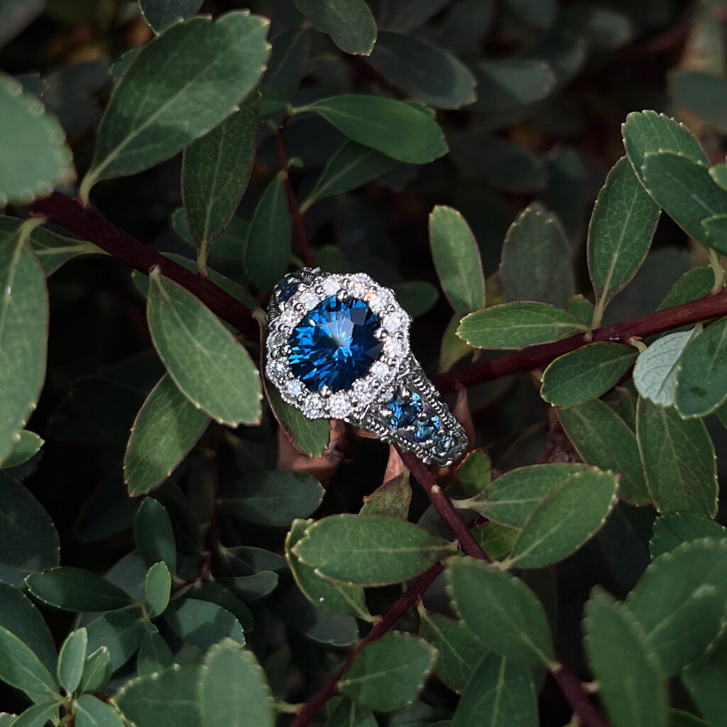 The 20240916_174409635_iOS copy sapphire and diamond ring displayed on lush green foliage.