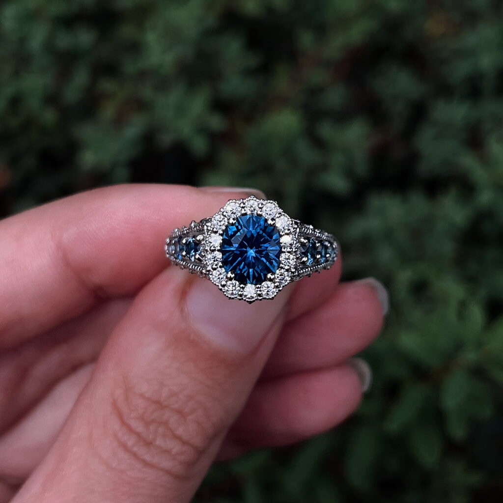 A hand displays the 20240916_174513540_iOS copy ring, which features a large blue center gemstone encircled by small clear stones and additional gems on the band, set against a blurred green foliage background.