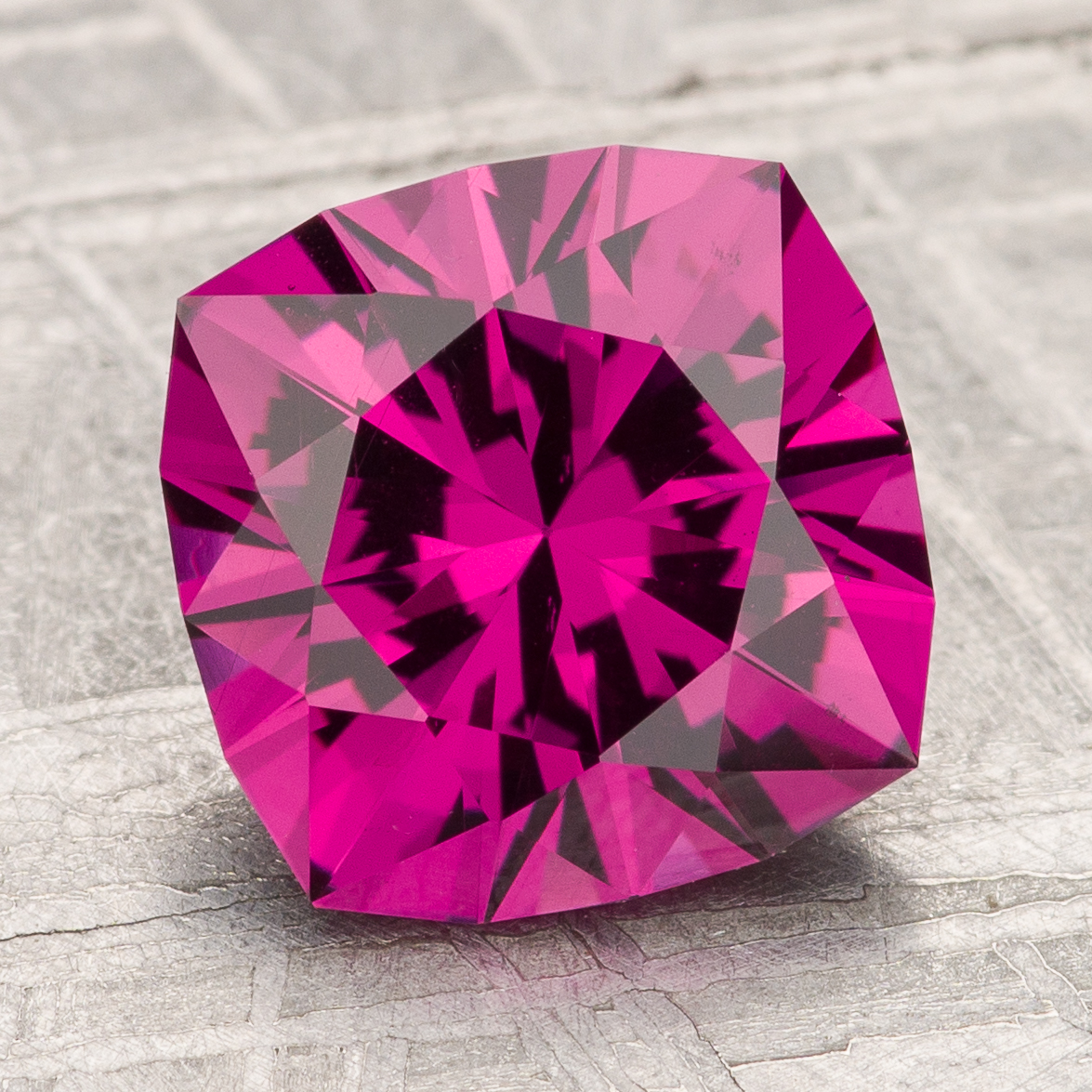 The Garnet-Magenta, a faceted cushion-cut gemstone in bright magenta, is displayed on a textured gray surface.
