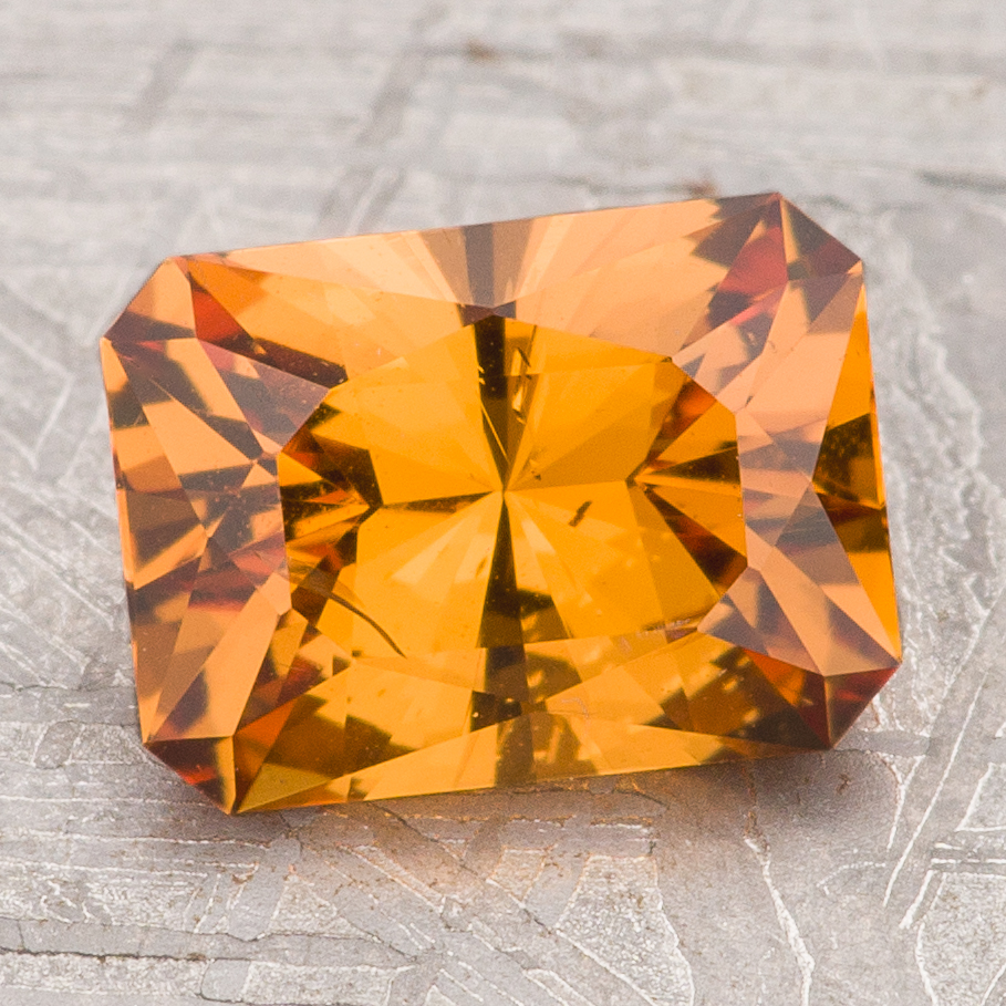 The Garnet-Orange gemstone features a rectangular, faceted brilliant cut and is displayed on a light gray textured surface.