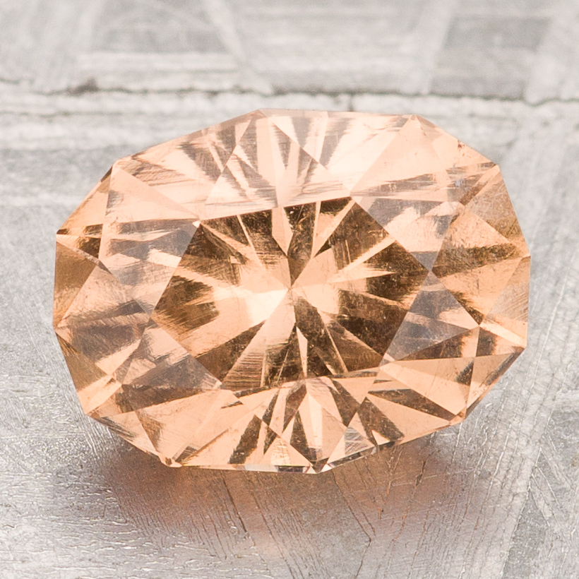 The Garnet-Peach, an oval-cut faceted gemstone with a peach hue, is displayed on a metallic textured surface.