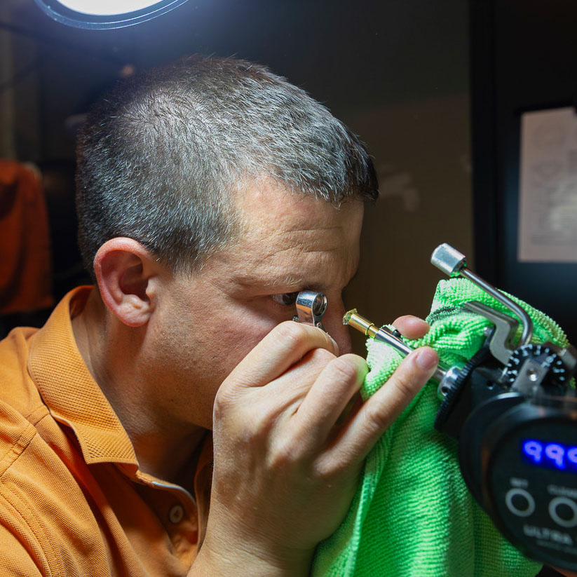 A man in an orange shirt examines "Our Story" from Earth's Treasury using a magnifying loupe, holding it with a green cloth under a bright light.