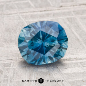 A 2.15-carat heated Montana sapphire is displayed on a textured light gray surface with "Earth's Treasury" at the bottom.