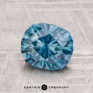 A 1.95-carat heated Montana sapphire with a geometric cut lies on a textured light gray surface. The text "EARTH'S TREASURY" appears at the bottom.