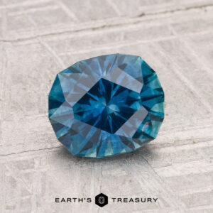 A 1.80-carat heated Montana sapphire, round brilliant-cut and blue, is displayed on a light textured surface with "Earth's Treasury" below.