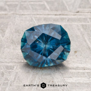 A faceted, oval 1.75-carat Montana Sapphire (heated) is showcased on a textured gray surface with "EARTH'S TREASURY" text at the bottom.