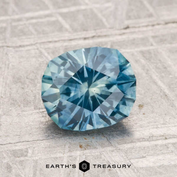 A 1.66-carat heated Montana sapphire, round brilliant-cut and blue, is shown on a faceted gray surface with "EARTH'S TREASURY" displayed underneath.