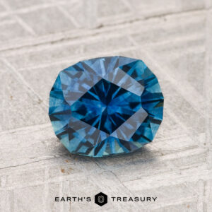 A 1.64-carat heated Montana sapphire with a cushion cut is showcased on a textured light gray surface, with "EARTH'S TREASURY" visible at the bottom.