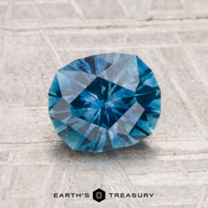 A 1.60-carat heated Montana sapphire is displayed on a textured light gray surface with the text "EARTH'S TREASURY" below it.