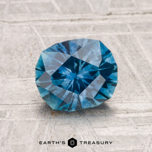 1.60-Carat Montana Sapphire (Heated) A 1.60-carat heated Montana sapphire is displayed on a textured light gray surface with the text "EARTH'S TREASURY" below it.