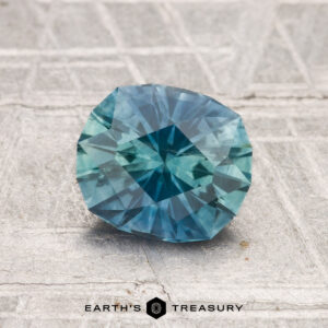 A 1.47-carat heated Montana sapphire with a blue-green hue is displayed on a textured gray surface, featuring the "EARTH'S TREASURY" text and logo at the bottom.