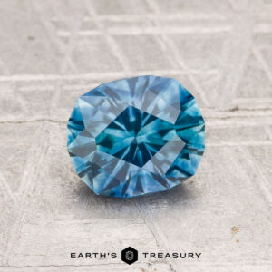 A 1.45-carat heated Montana sapphire with blue geometric facets rests on a textured gray surface. The Earth's Treasury logo appears at the bottom of the image.