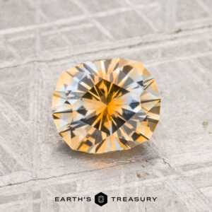 A 1.40-carat heated Montana sapphire with a brilliant cut is displayed on a textured gray surface. The "EARTH'S TREASURY" logo appears at the bottom.