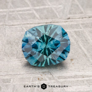A 1.39-carat heated Montana sapphire with a blue-green hue is displayed on a textured gray surface, with "EARTH'S TREASURY" text at the bottom.