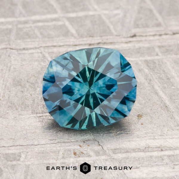 1.39-Carat Montana Sapphire (Heated) A 1.39-carat heated Montana sapphire with a blue-green hue is displayed on a textured gray surface, with "EARTH'S TREASURY" text at the bottom.