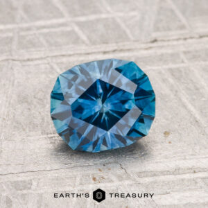 A 1.38-carat heated Montana sapphire is showcased on a textured light gray surface with the text "EARTH'S TREASURY" below it.