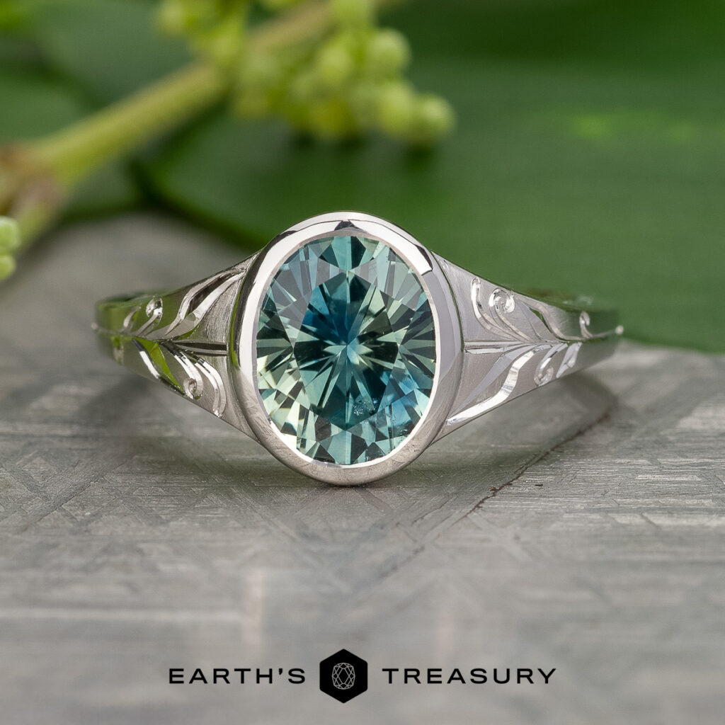 The _SNY0193-Edit-1200-BW ring features an oval blue-green gemstone set in silver, displayed on a textured surface with green leaves in the background. "EARTH'S TREASURY" is shown at the bottom.