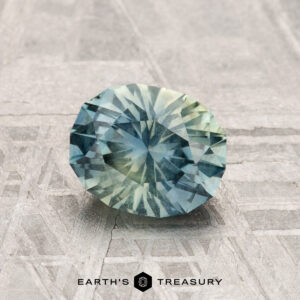 A round, faceted 1.30-carat Montana sapphire in blue-green sits on a patterned gray surface with "EARTH'S TREASURY" at the bottom.