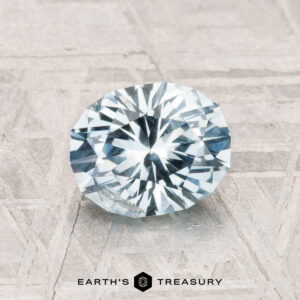 A 1.30-carat Montana Sapphire with a round brilliant cut and pale blue hue rests on a textured gray surface. “EARTH'S TREASURY” appears at the bottom.