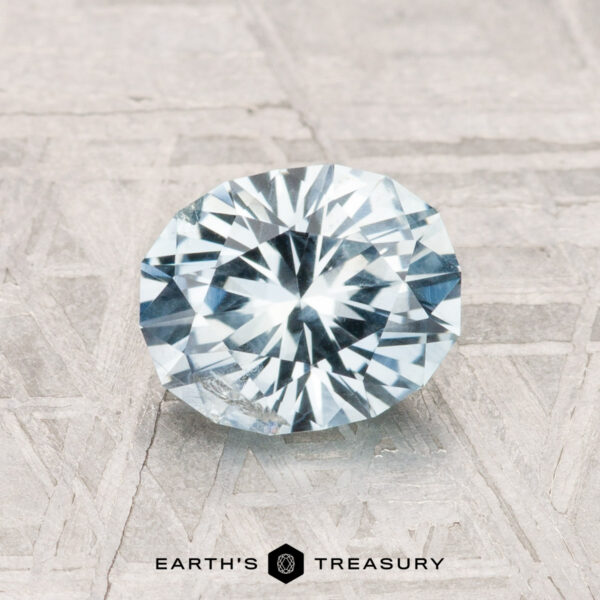 1.30-Carat Montana Sapphire A 1.30-carat Montana Sapphire with a round brilliant cut and pale blue hue rests on a textured gray surface. “EARTH'S TREASURY” appears at the bottom.