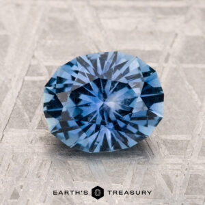 A round, brilliantly cut 2.03-carat Montana Sapphire in medium blue is displayed on a geometric patterned surface, with the text "EARTH'S TREASURY" at the bottom.