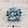A stunning 1.75-Carat Montana Sapphire in round brilliant-cut blue-green hues rests on a textured metallic surface, with the "EARTH'S TREASURY" logo displayed beneath.
