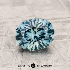 A stunning 1.75-Carat Montana Sapphire in round brilliant-cut blue-green hues rests on a textured metallic surface, with the "EARTH'S TREASURY" logo displayed beneath.