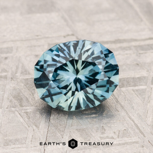1.75-Carat Montana Sapphire A stunning 1.75-Carat Montana Sapphire in round brilliant-cut blue-green hues rests on a textured metallic surface, with the "EARTH'S TREASURY" logo displayed beneath.