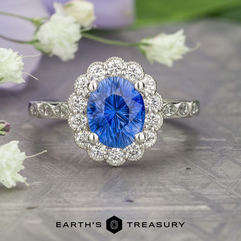 The _SNY0373-Edit-1200-BW is a silver ring featuring an oval blue gemstone encircled by small round diamonds, showcased on a textured surface with white flowers in the background.