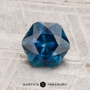 A 1.53-carat heated Montana sapphire with an octagonal shape is displayed on a light textured surface, with the text "EARTH'S TREASURY" below.