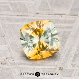 A 1.46-carat heated Montana sapphire with a yellow-green gradient, cushion-cut and displayed on a textured surface, features "Earth's Treasury" branding at the bottom.