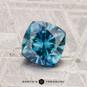 A 1.40-carat heated Montana sapphire with a cushion cut rests on a textured gray surface, with "EARTH'S TREASURY" displayed at the bottom.