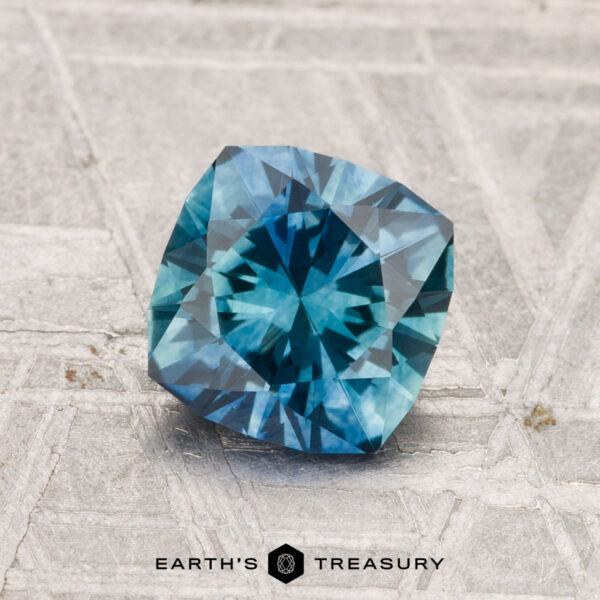 1.40-Carat Montana Sapphire (Heated) A 1.40-carat heated Montana sapphire with a cushion cut rests on a textured gray surface, with "EARTH'S TREASURY" displayed at the bottom.