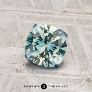 A cushion-cut 1.05-carat heated Montana sapphire in pale blue-green rests on a textured light gray surface featuring the "Earth's Treasury" logo at the bottom.