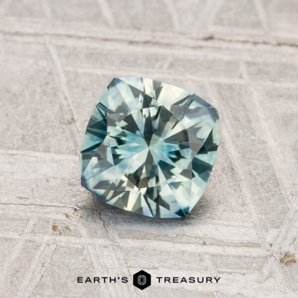 1.05-Carat Montana Sapphire (Heated) A cushion-cut 1.05-carat heated Montana sapphire in pale blue-green rests on a textured light gray surface featuring the "Earth's Treasury" logo at the bottom.